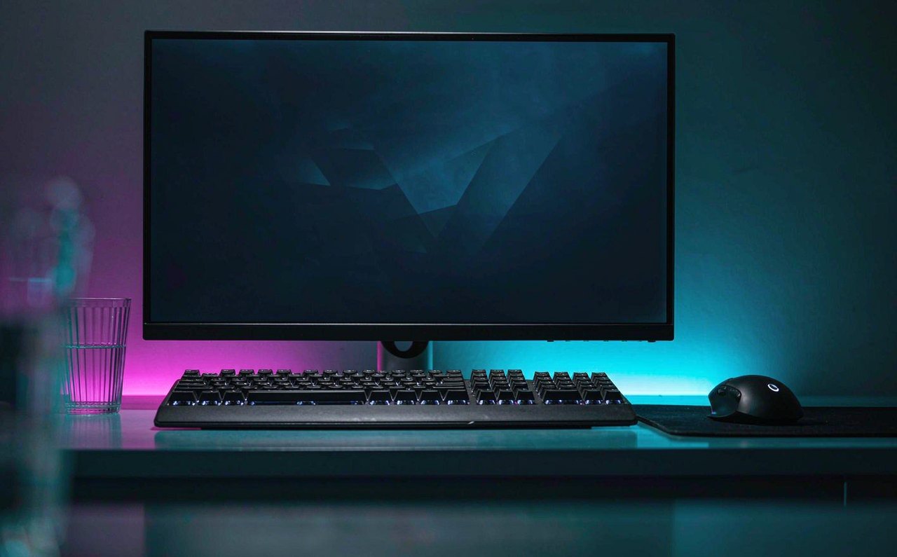 A dark gaming desk at night with a monitor glowing in blue and magenta ambient light