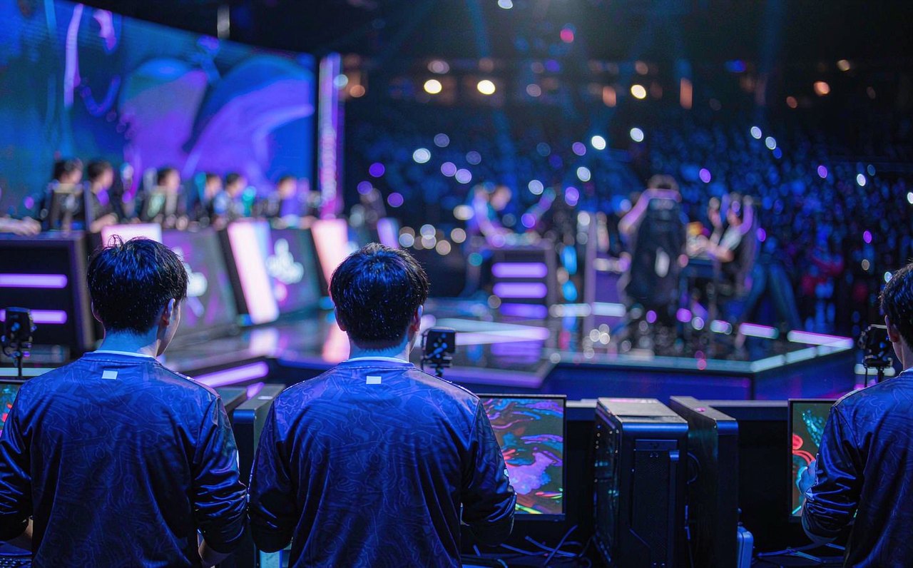Professional esports players seen from behind at competition PCs on a lit arena stage, with blurred audience lights in the background