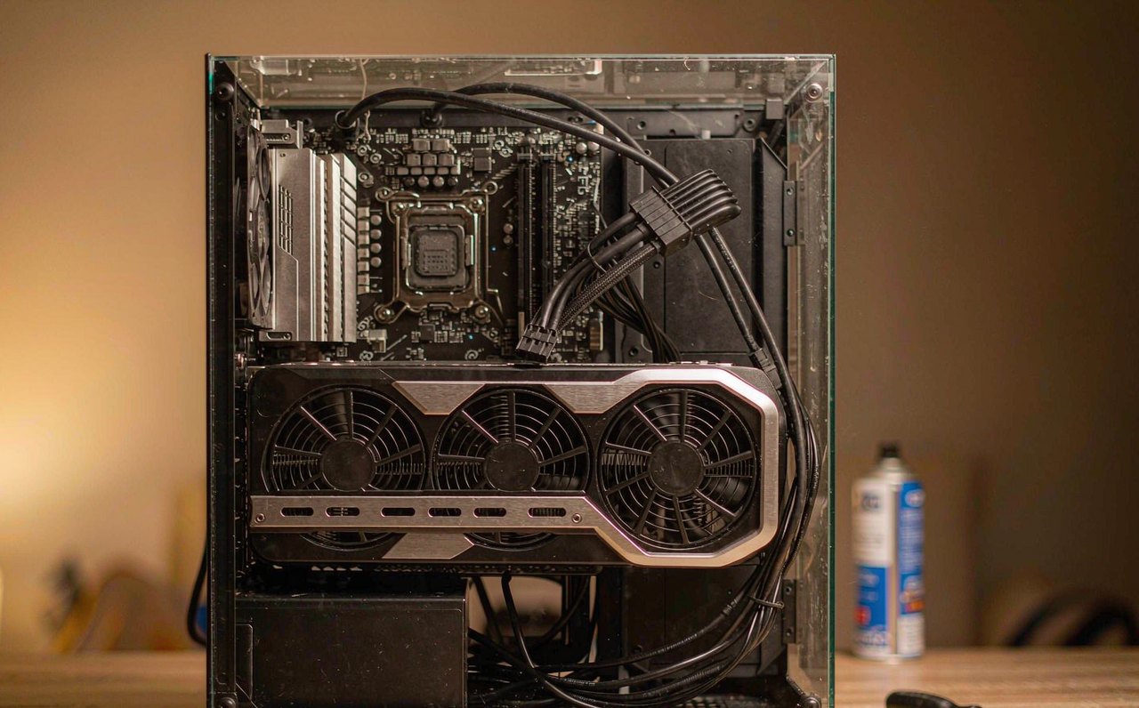 A desktop gaming PC with its side panel removed, showing a large graphics card lit by warm indoor light on a wooden desk