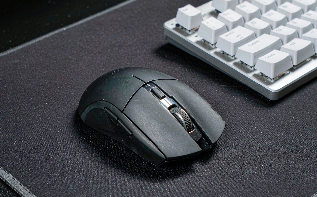 A lightweight gaming mouse photographed on a large dark mousepad next to a mechanical keyboard, studio lighting